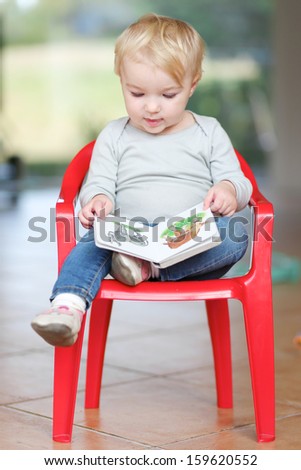Lovely little baby girl sitting indoors on a small red plastic chair reading children book about animals