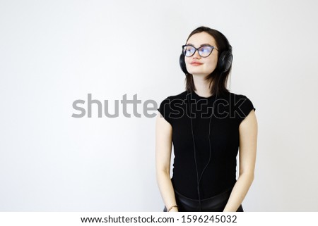 Satisfied girl holds hands on headphones, enjoys loud sound, listens favourite track, opens mouth while sings, wears black t shirt, stands over white background with free space