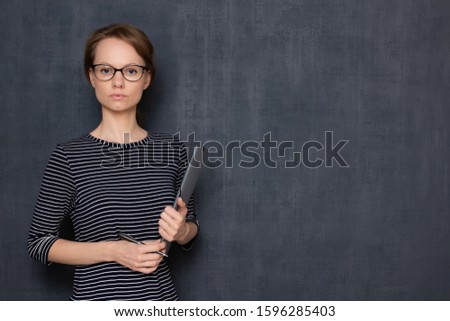 Studio portrait of serious focused young woman with glasses, wearing jumper, looking attentively and strictly at camera, holding folder and pen in hands, over gray background, copy space on right