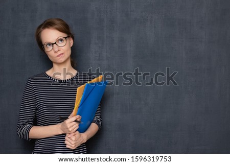 Studio waist-up portrait of thoughtful focused fair-haired girl with glasses, wearing striped jumper, looking aside, holding colored folders and pen in hands, over gray background, copy space on right