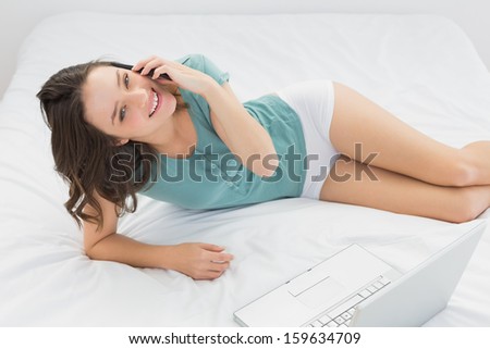 Portrait of a casual young woman using mobile phone by laptop in bed at home