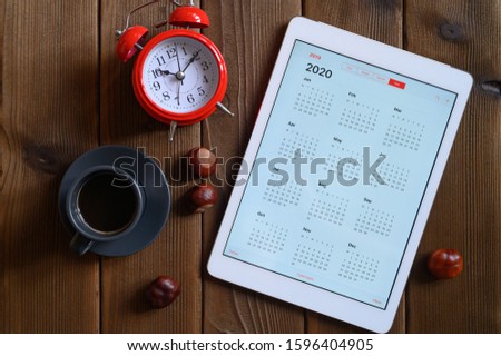 a tablet with an open calendar for 2020, a Cup of coffee, chestnuts and a red alarm clock on a wooden background