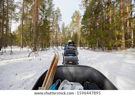 People riding a snowmobile, towing a trailer on the background of a snowy forest in winter in sunny weather. From the sled stick ski