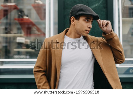 Handsome amazing young arabian or egyptian guy posing on camera. Stand and look to right. Wear brown jacket and grey cap. Model alone on street outside. Isolated on multicolored background