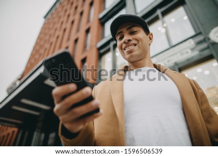 Low view of arabian or egyptian young man hold black smartphone in hand. Look at it and smile. Phone user. Modern technologies. Stand near urban orange building. Electronics