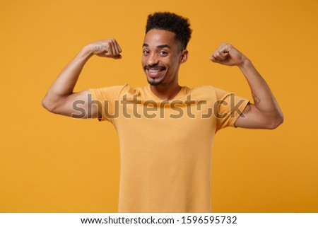 Smiling young african american guy in casual t-shirt posing isolated on yellow orange background studio portrait. People sincere emotions lifestyle concept. Mock up copy space. Showing biceps muscles