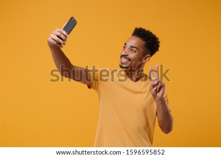 Smiling young african american guy in casual t-shirt posing isolated on yellow orange background. People lifestyle concept. Mock up copy space. Doing selfie shot on mobile phone showing victory sign