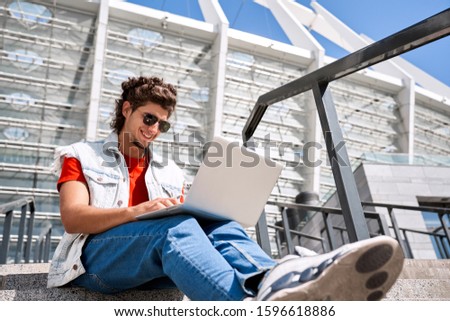 Young man wearing sleeveless jean jaket and sunglasses sitting on stairs outdoors spending free time browsing internet on laptop smiling happy bottom view