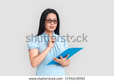 Asian woman wearing eyeglasses standing isolated on white background holding clipboard and pen looking aside thoughtful