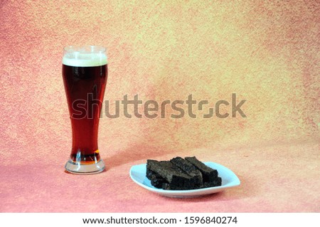 A tall glass of dark beer with white foam and a plate with fresh wheat croutons on a brown background. Close-up.
