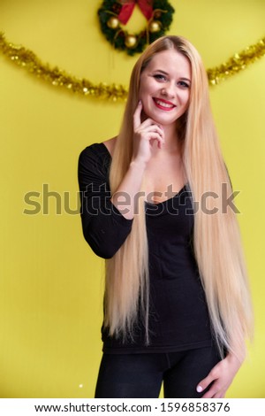 Concept of a young blonde woman with New Year's decor. Portrait of a cute girl in a black T-shirt with long beautiful hair and great makeup. Smiling, showing emotions on a yellow background.