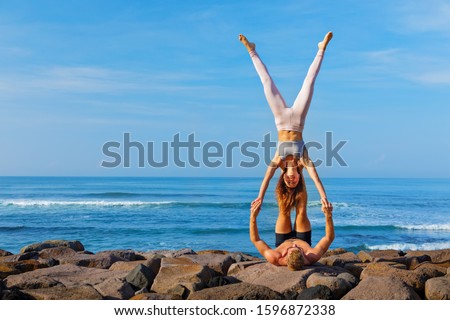 Fit young couple doing acro yoga at spa retreat on sea beach. Active woman on partner feet, balancing at handstand acroyoga pose. Healthy lifestyle. People outdoor sport activity on family vacation.