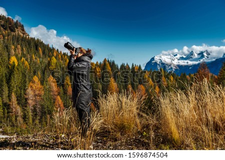 Young female photographer dressed in black jacket taking pictures of dolomites in Italian alps /  Dolomite mountains in background during sunny day / High ISO image