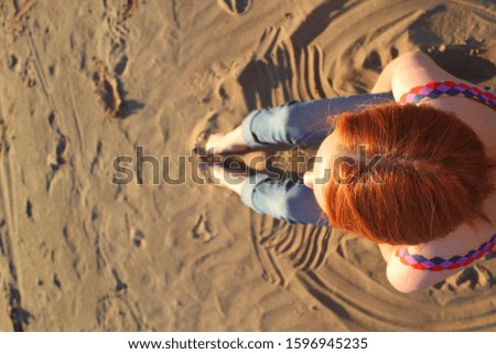 Beautiful girl with bright red hair lit by the sun sits on the sand. Summer mood. Stock photo with empty space for your text. No face, top view