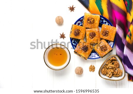 Traditional arabic dessert baklava with cashew, walnuts, raisins on plate, bowl, teapot with Uzbek national ornament on white wooden table. Homemade baklava with nuts and honey Top view Flat Lay