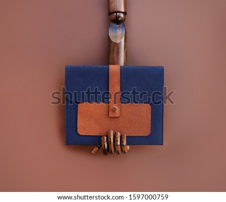 A wooden fashion dummy hand holding a dark blue felt and leather clutch, on brown background