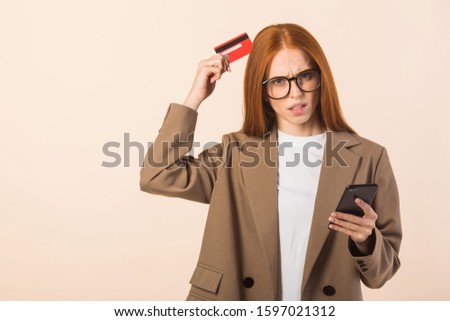 beautiful young woman with red hair in a jacket on a beige background with a credit card and a phone in her hands