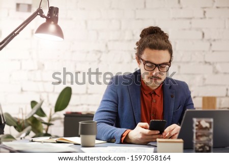 Young businessman in eyeglasses sitting at his workplace and using mobile phone he working at office