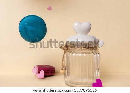 A glass can with heart-shaped lid, blue flying macaroon are on a light background. Close-up, layout design.