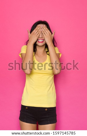 Surprised young woman in yellow shirt and black shorts covers her eyes with hands and smiles. Three quarter length studio shot on pink background.
