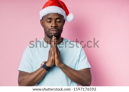Young religious calm african american man wearing white t shirt and Santa hat, keeping palms together, prying with closed eyes , hoping for best, asking for a miracle on Christmas. Meditation, faith.