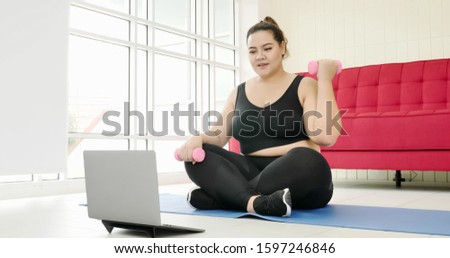 Chubby woman doing exercise by dumbbell and surfing internet in a room.