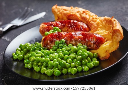 close-up of a portion of Toad in the hole, Sausage Toad, sausages in Yorkshire pudding batter on a black plate served with green peas and onion gravy,  view from above