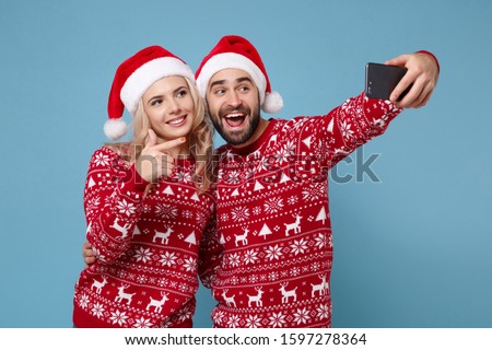 Funny young couple guy girl in Christmas sweater Santa hat posing isolated on blue background. New Year 2020 celebration holiday party concept. Mock up copy space. Doing selfie shot on mobile phone
