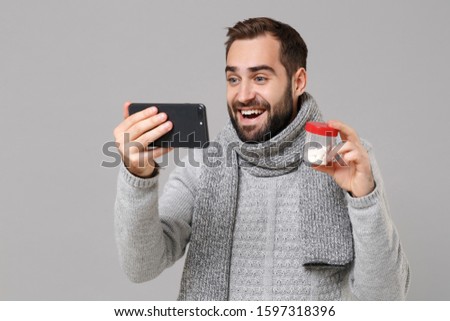 Funny young man in gray sweater, scarf isolated on grey background. Healthy lifestyle ill sick disease treatment, cold season concept. Hold tablets doing selfie shot on mobile phone making video call