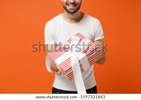 Cropped image of man in white t-shirt posing isolated on orange background. Valentine's Day Women's Day birthday holiday concept. Mock up copy space. Hold red striped present box with gift ribbon bow