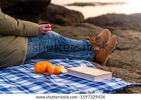 Girl on the beach on vacation on a plaid drinks tea, a book and tangerines in the front pallet, autumn mood