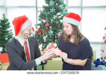 Male and female ethnicity Russian holding gift box presenting the gift box. Both look at the face and smile on Christmas Eve or New Year Day The backdrop has a Christmas tree and the gift pile.