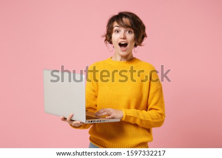 Shocked young brunette woman girl in yellow sweater posing isolated on pastel pink background studio portrait. People lifestyle concept. Mock up copy space. Holding and working on laptop pc computer