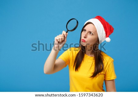 Serious young woman Santa girl in yellow t-shirt Christmas hat posing isolated on blue wall background. Happy New Year 2020 celebration holiday concept. Mock up copy space. Holding magnifying glass