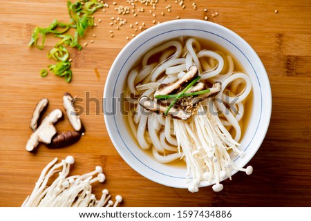 Japanese noodles. Udon noodles. Noodles with enoki mushrooms served in a bowl placed atop a wooden cutting board. Classic Japanese cuisine udon noodles with a hearty beef broth and enoki mushrooms. 