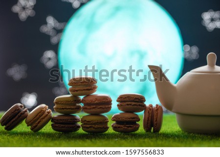Tea time.Teapot with tea, on green grass against the background of the moon and the starry sky, with french sweets macaroons. East picture. Delicious tea