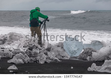 Photographer standing on black sand take pictures the waves. Melting glacier ice In the foreground. Iceland