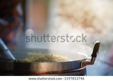 Cooking person in the restaurant is cooking while using a sieve for scalding yellow noodles in a large pot. The water is boiling and mass of steam reflected in the morning light.