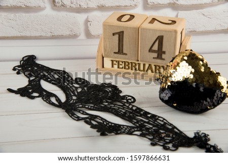14 february on wooden cube calendar. Saint Valentine's holiday decorations. Soft toy heart with golden sequins, black lace eye carnival mask. Love, romance greetings