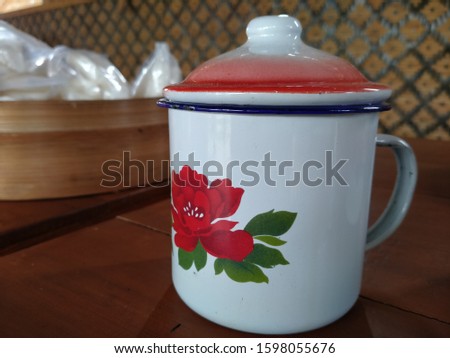 The enamel cup with the lid and flower motif, Bamboo tray and wicker wall as background.