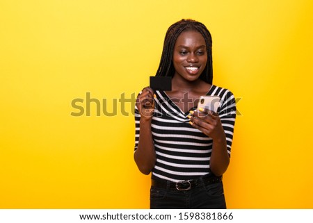 Picture of excited happy african woman posing isolated over yellow background using mobile phone holding credit card.