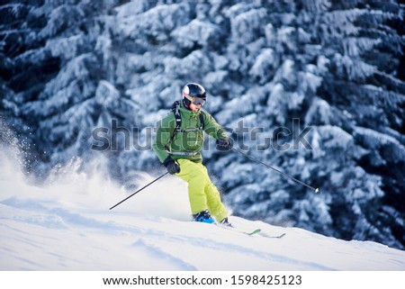 Proficient skier coming down along wooded hillside using professional ski equipment and making deep snow powder. Winter outdoors activities concept. Picturesque forest scenery on background. Side view Royalty-Free Stock Photo #1598425123