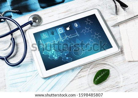 Laboratory patient examination in doctors office. Tablet computer with medical app interface on screen. Stethoscope, x-ray image and cardiogram on wooden desk. Medical diagnostics and examination