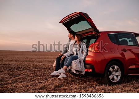 Young Happy Couple Dressed Alike in White Shirt and Jeans Sitting at Their New Car Trunk, Beautiful Sunset on the Field, Vacation and Travel Concept Royalty-Free Stock Photo #1598486656