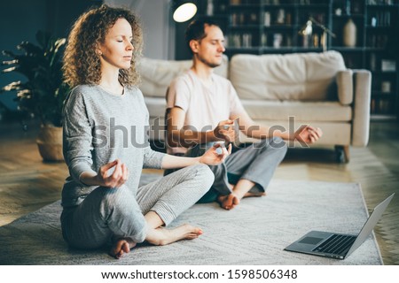 Calm couple in pajamas meditating, listening spiritual practices lessons on laptop, sitting on lotus pose at home. Yoga concept. Royalty-Free Stock Photo #1598506348