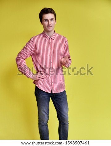 Vertical portrait of a handsome young man with a short haircut with a broad smile in a pink shirt and jeans on a yellow background. Cute looks at the camera, standing straight.