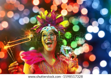 Beautiful young woman in carnival mask and stylish masquerade costume with feathers and sparklers in colorful bokeh on black background. Christmas, New Year, celebration. Festive time, dance, party.