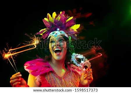 Beautiful young woman in carnival mask and stylish masquerade costume with feathers and sparklers in colorful lights on black background. Christmas, New Year, celebration. Festive time, dance, party.