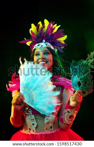 Beautiful young woman in carnival mask and stylish masquerade costume with feathers fan in colorful lights and glow on black background. Christmas, New Year, celebration. Festive time, dance, party.