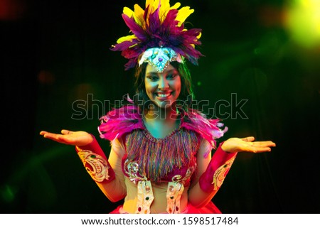 Beautiful young woman in carnival mask and stylish masquerade costume with feathers in colorful lights and glow on black background. Christmas, New Year, celebration. Festive time, dance, party.